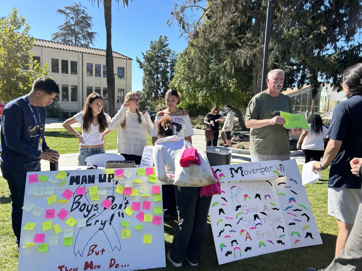 Students learn about men’s health with posters encouraging open discussion on topics like mental health. Colorful sticky notes and mustache decorations highlight messages on masculinity and support for the cause.